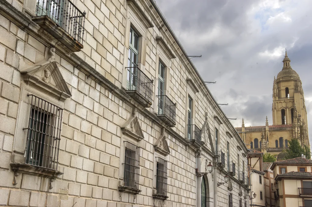 Vista de un detalle de la fachada del Palacio Episcopal de Segovia con un gran portón verde y la torre de la Catedral de Segovia al fondo.