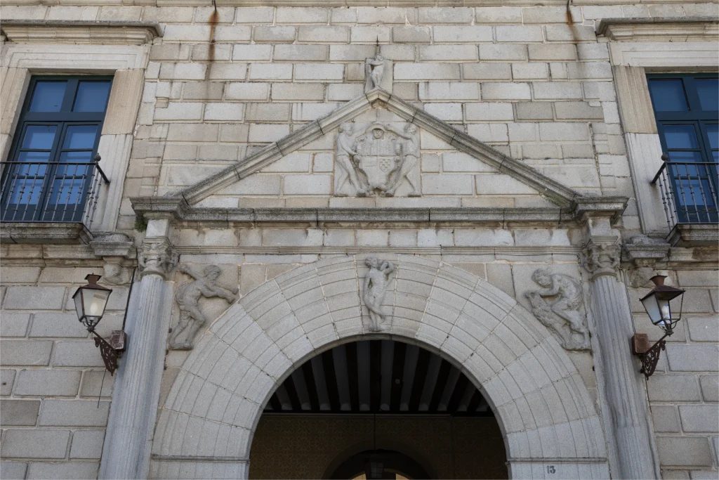 Fachada del Palacio Episcopal de Segovia con un arco de medio punto decorado con esculturas y relieves, ventanas con marcos de piedra y balcones de hierro forjado.